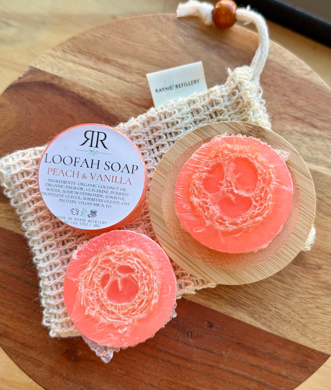 Pink loofah soap discs with 'Peach & Vanilla' scent label on wooden tray with woven pouch