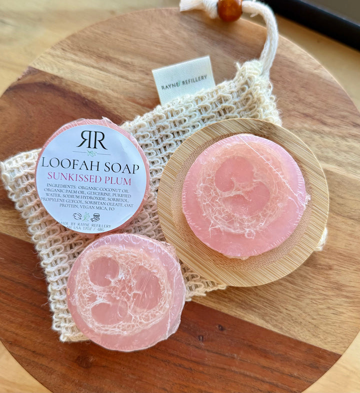 Pink loofah soap discs labeled 'Sunkissed Plum' on wooden tray with woven soap bag