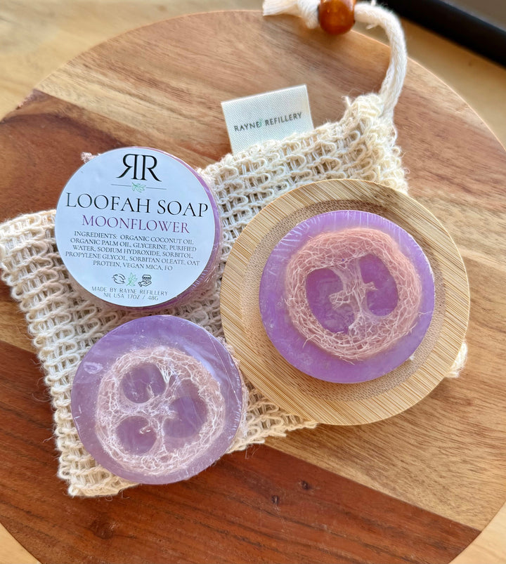 Purple loofah soap bar with flower cutout, round bar on wooden board and mesh bag