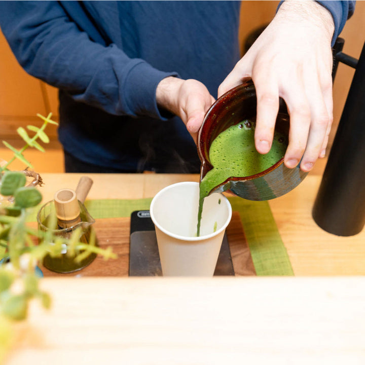 Pouring green matcha from a small pitcher into a cup on a tea tray