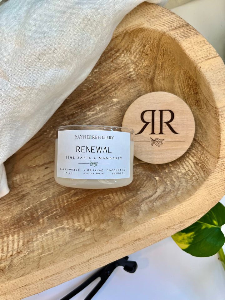 White glass coconut soy candle labeled RENEWAL with round wooden lid