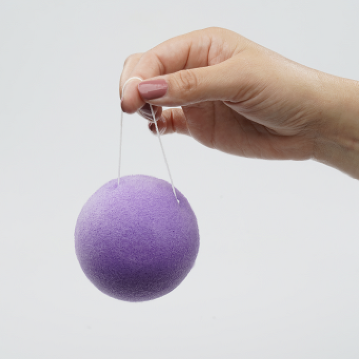 Purple konjac facial cleansing sponge disc with string, held above skin
