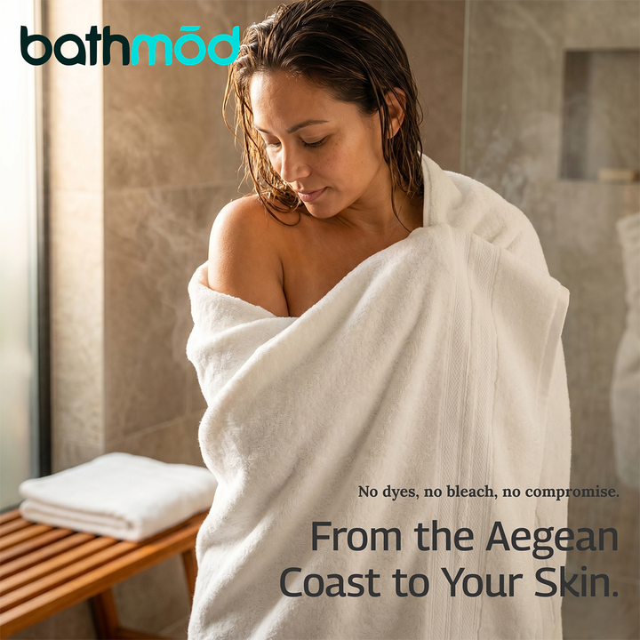 White bath towel wrap on a person in a bathroom; stacked towels visible in the background.