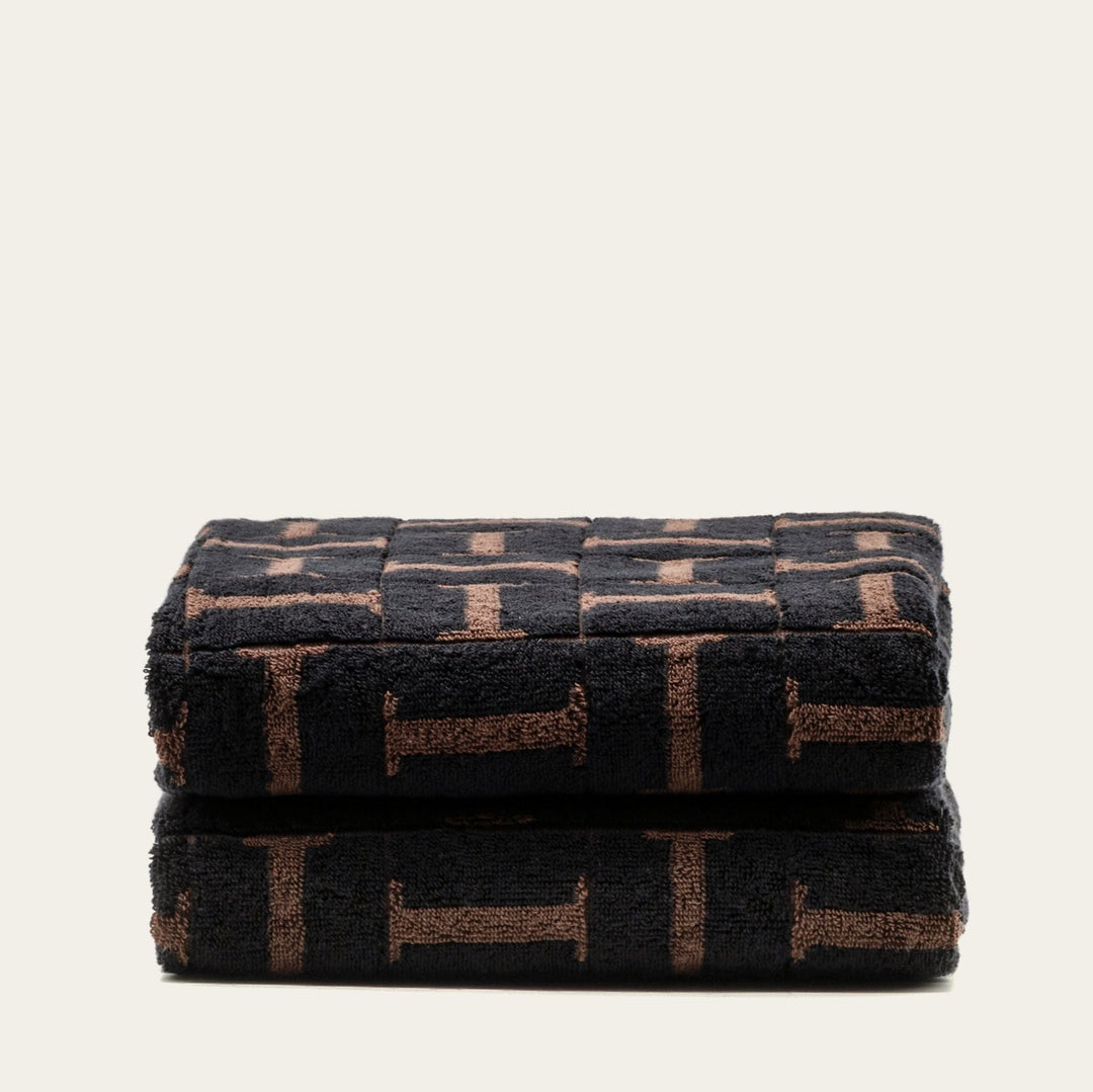 Folded bath towel with dark base and tobacco-brown block motif, stacked neatly.