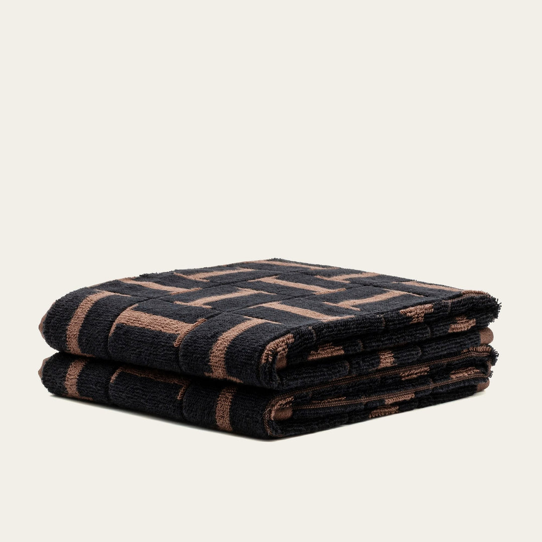 Folded towel with dark fabric and tobacco-brown geometric lines, plush cotton.