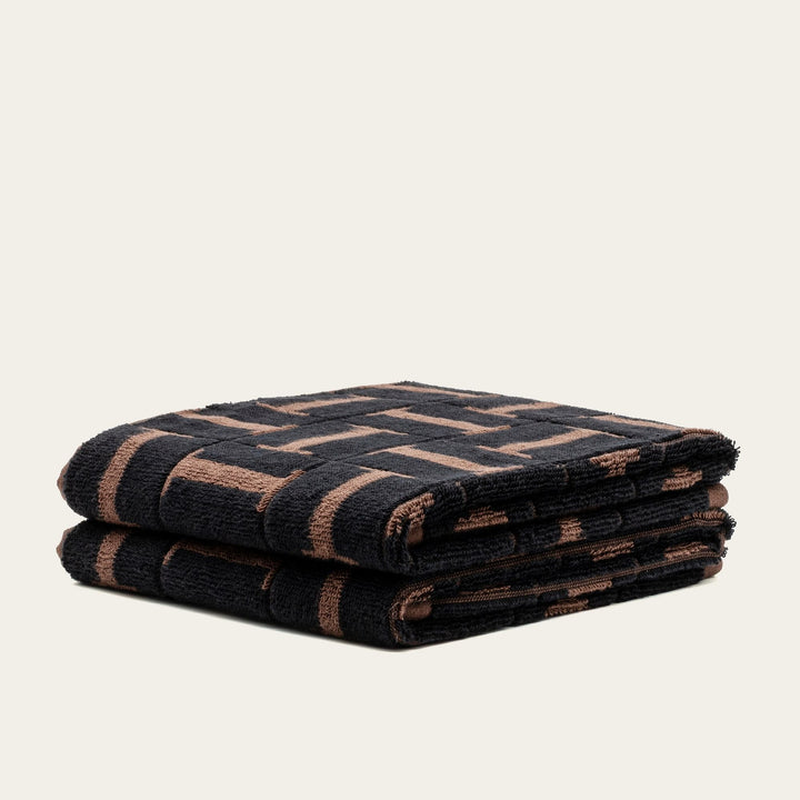 Folded towel with dark fabric and tobacco-brown geometric lines, plush cotton.
