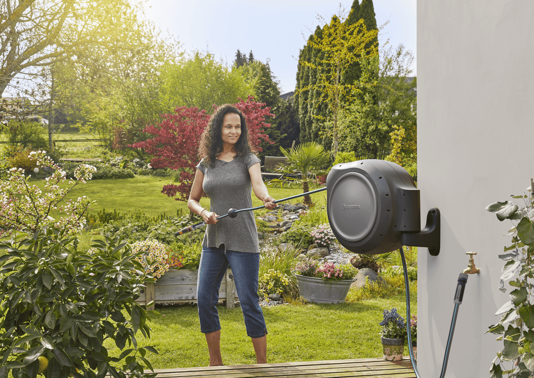 Retractable garden hose reel on wall with gray drum, person spraying flowers