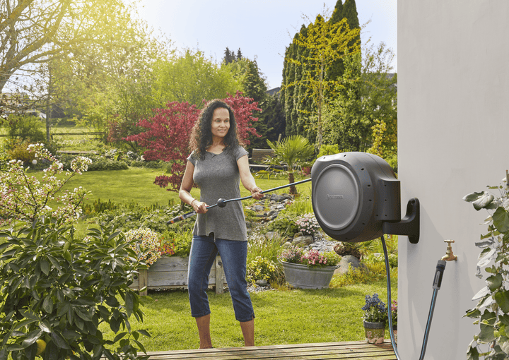 Retractable garden hose reel on wall with gray drum, person spraying flowers