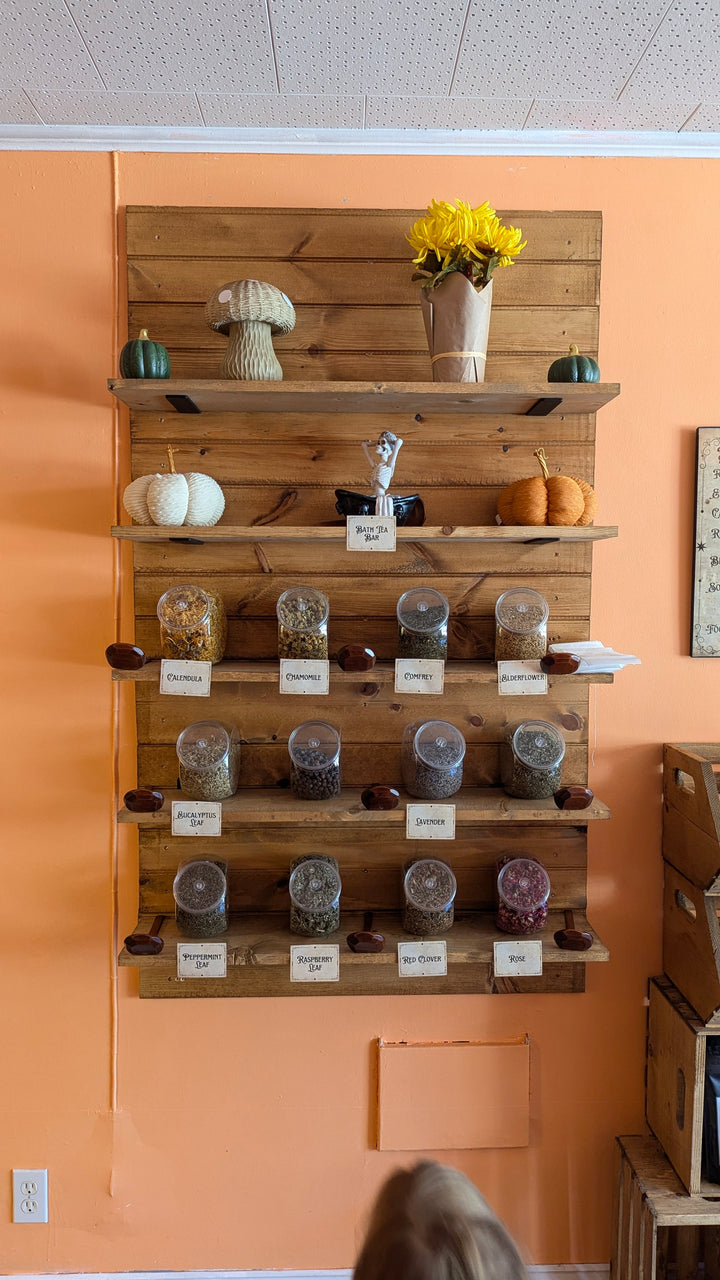 Wooden display shelf of labeled bath tea jars, with small bottles and scoops.
