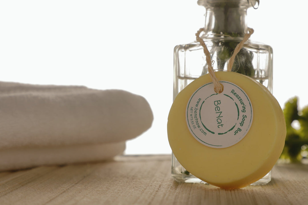 Round pale yellow soap bar with paper label tag, near a glass bottle on a wooden surface.