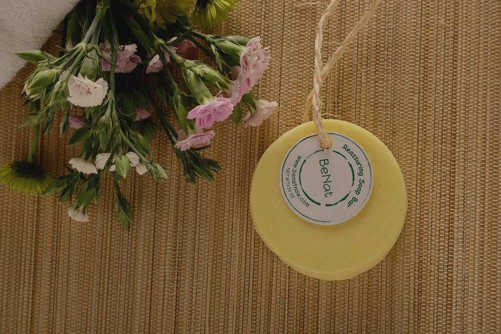 Round pale yellow soap bar with label tag on woven mat, near a small flower bouquet.