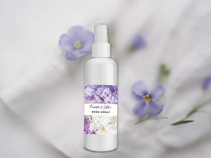 Room spray bottle with white pump on white fabric beside purple flowers