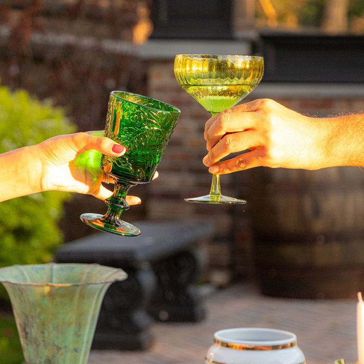 Ribbed sage-green coupe glasses clinking in outdoor setting, hands holding the stems.