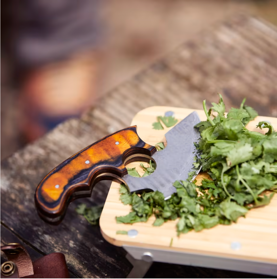 Ulu-style blade with orange wood handle and leather sheath beside herbs on board