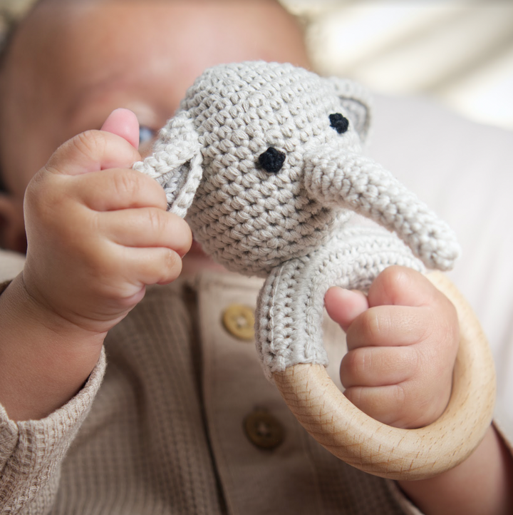 Grey crochet elephant rattle teether with black eyes and wooden ring, held by baby