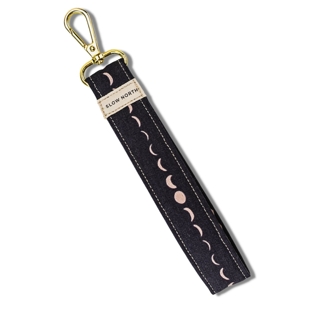 Dark fabric wristlet keychain with gold clasp, crescent moon stitch pattern, SLOW NORTH label.