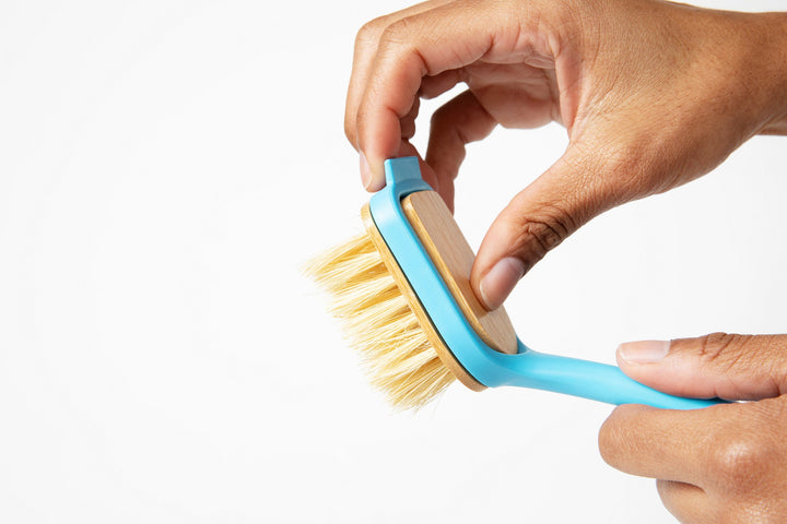 Hand gripping dish brush with blue handle and yellow bristle brush head
