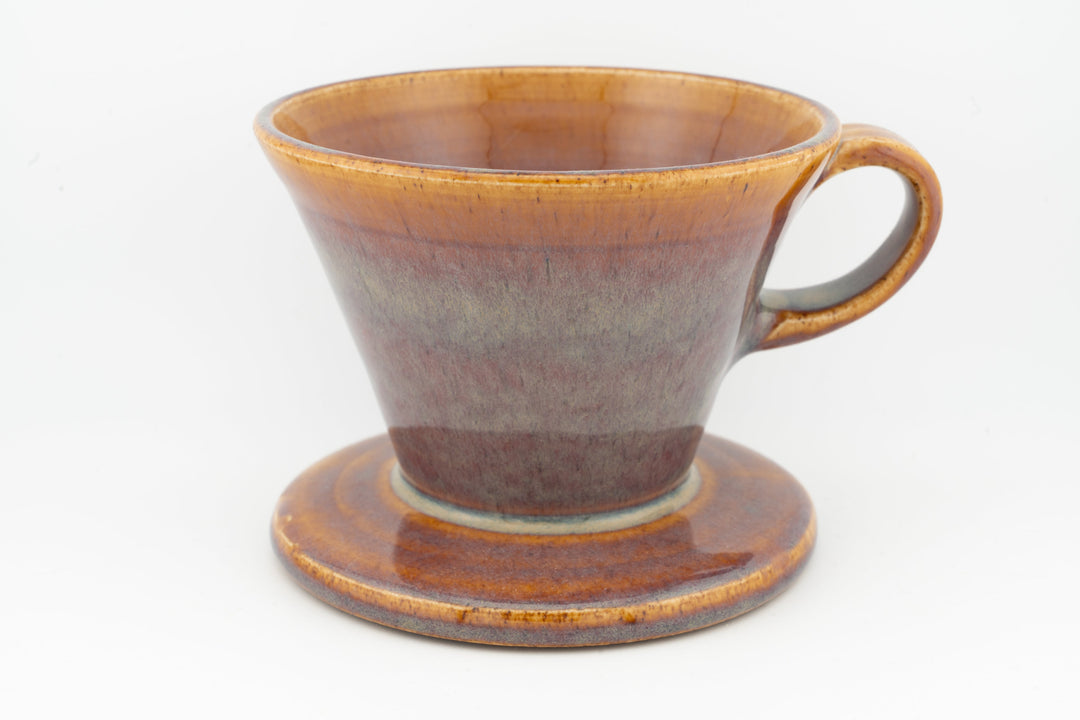 V60 ceramic coffee dripper with drip stand, amber-brown glaze and side handle.