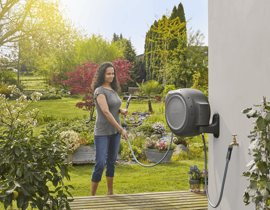 Wall-mounted retractable hose reel with gray housing, hose spraying near garden beds