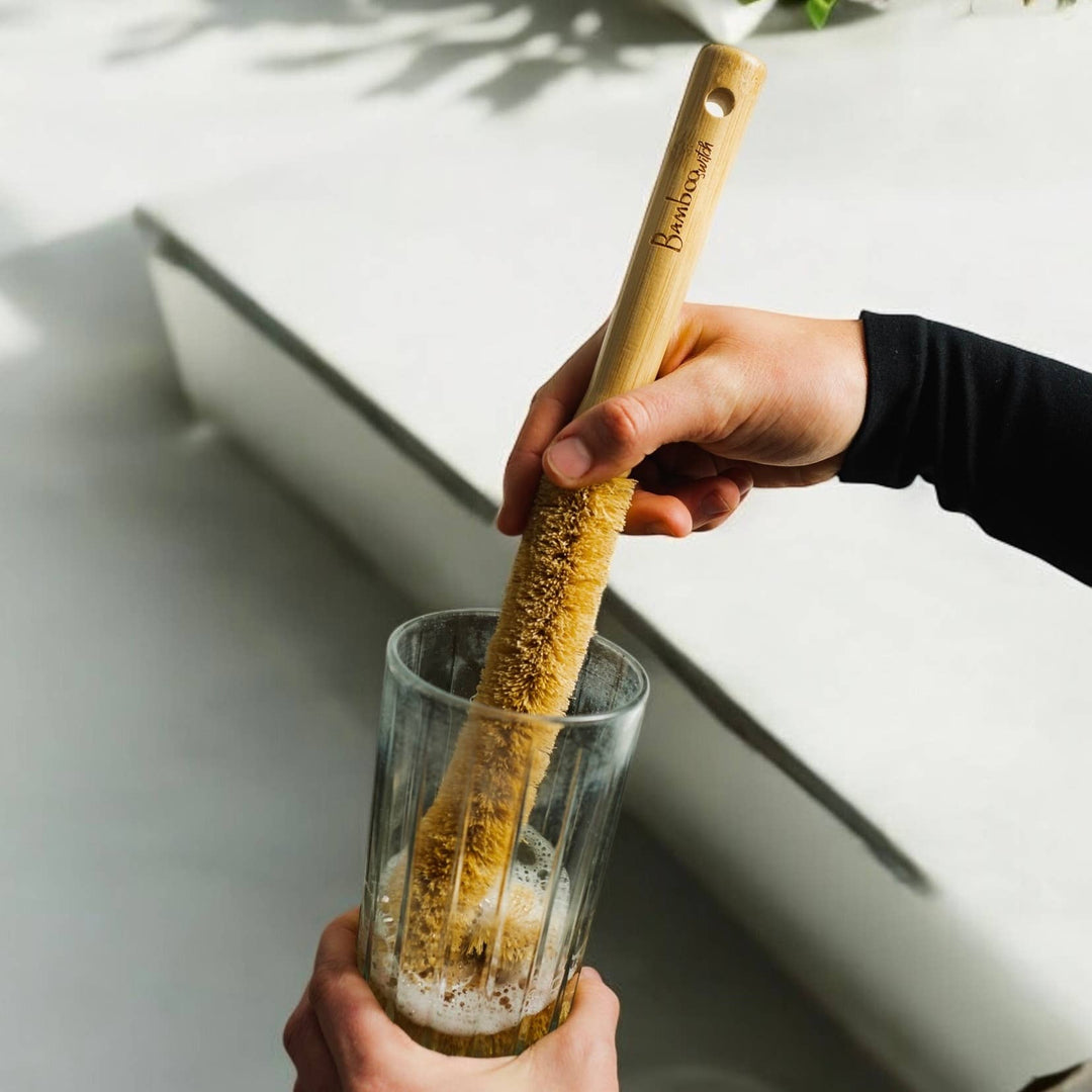 Bottle brush being used in a clear glass with yellow-brown bristles and wooden handle