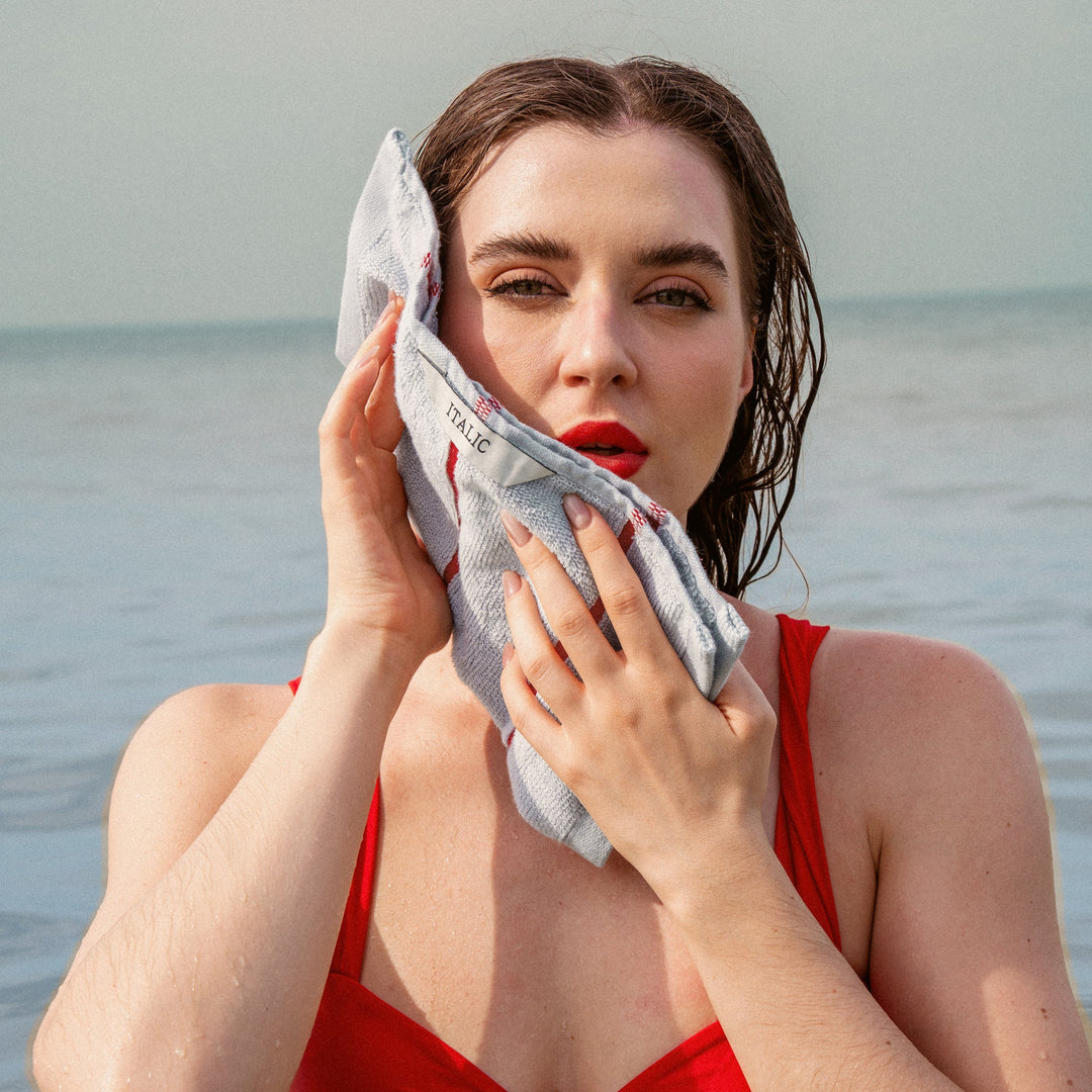 Gray towel with red striped border held in hands near the ocean, for face drying.