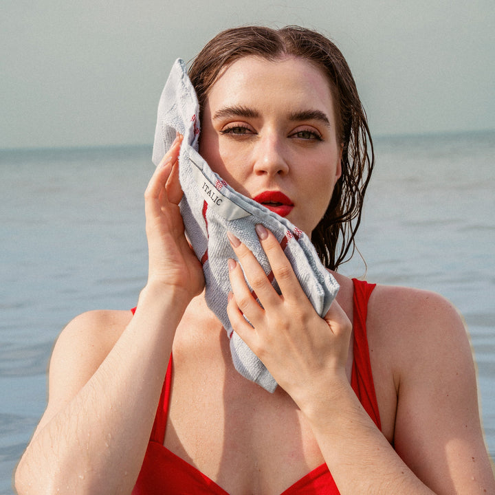 Gray towel with red striped border held in hands near the ocean, for face drying.