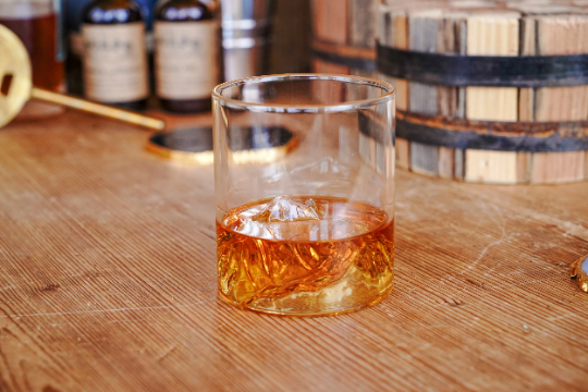 Clear whiskey tumbler with amber whiskey, ice cubes, and mountain range etching