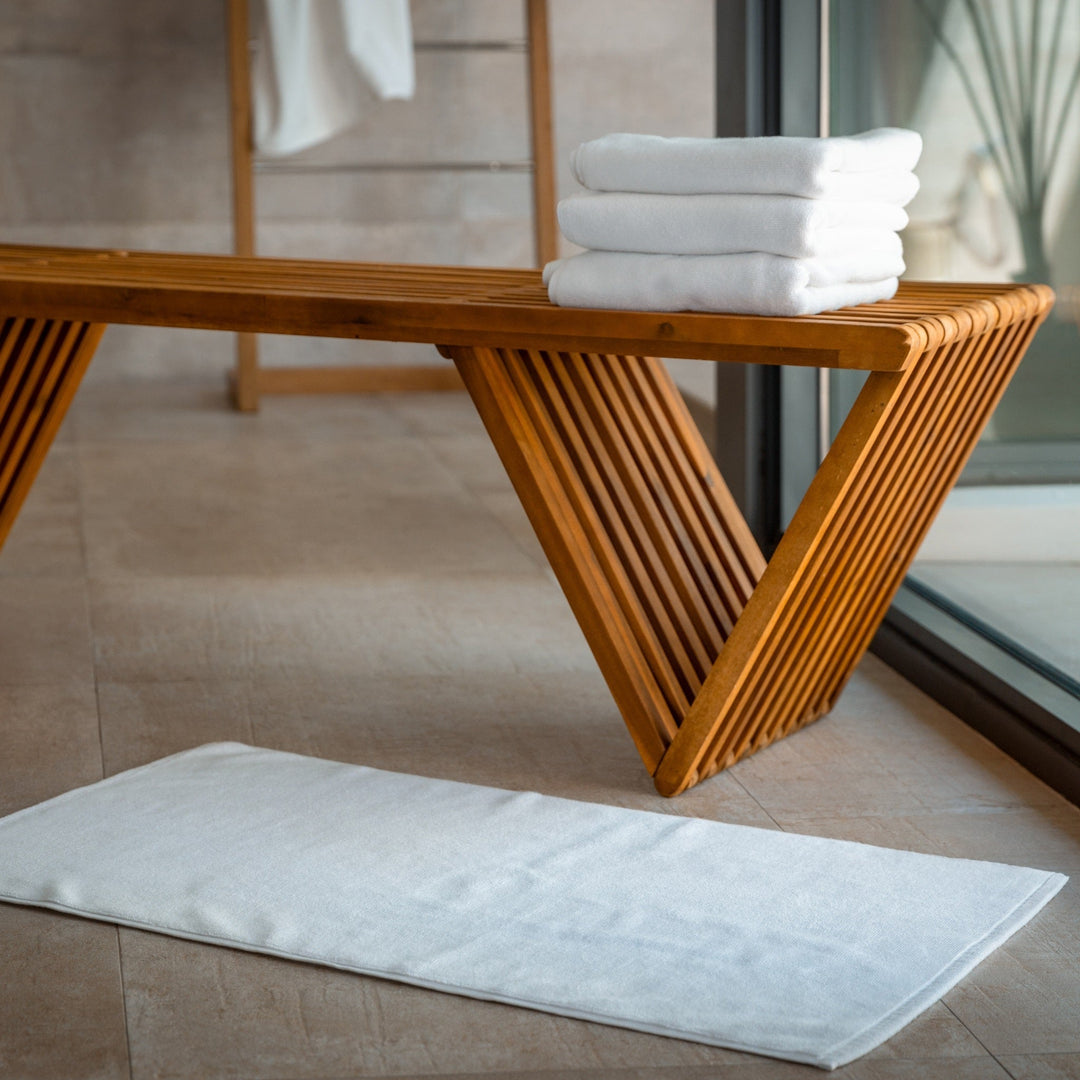 White bath mat laid flat on the floor beside a wooden bench and stacked towels.