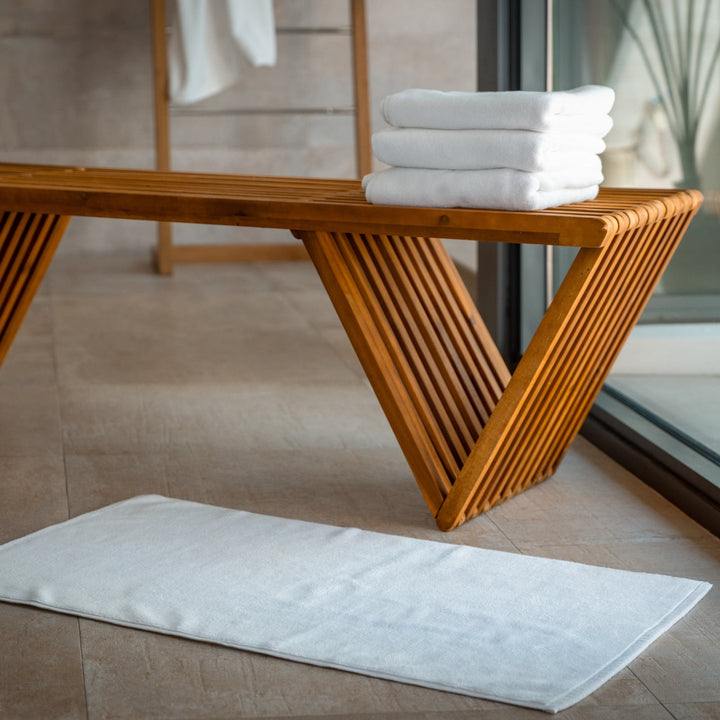 White bath mat laid flat on the floor beside a wooden bench and stacked towels.