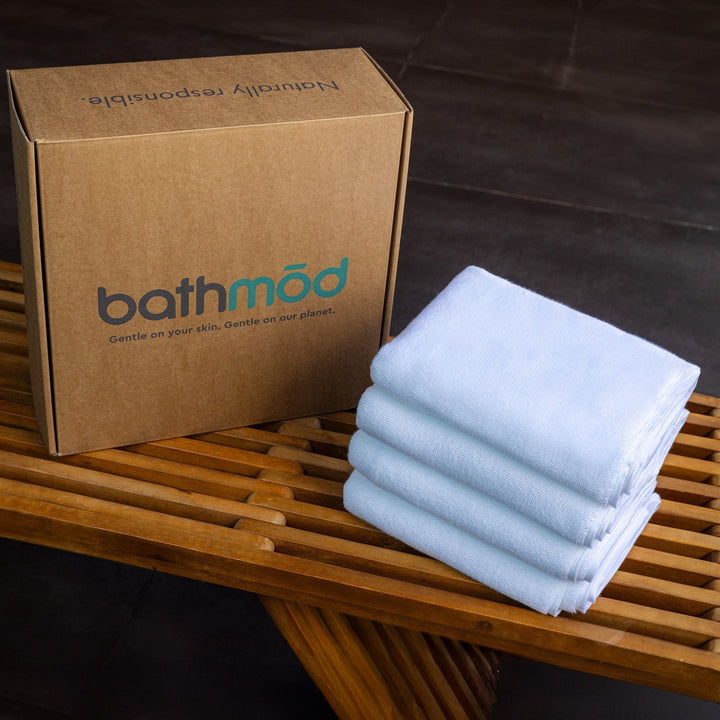White bath mats folded in a stack beside a branded gift box on a wooden rack.