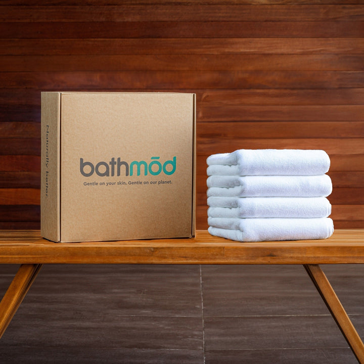 Four-pack white bath mats with a branded cardboard box on a wooden bench.