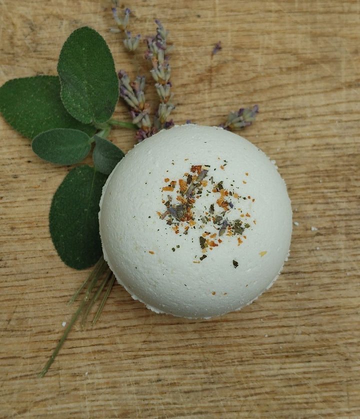 White bath bomb with orange and green specks, with dried herb sprigs nearby