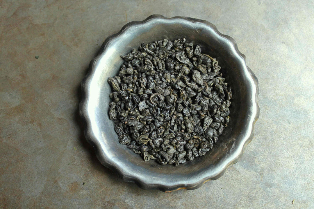 Loose green tea leaves in a metal bowl, dark curled leaves
