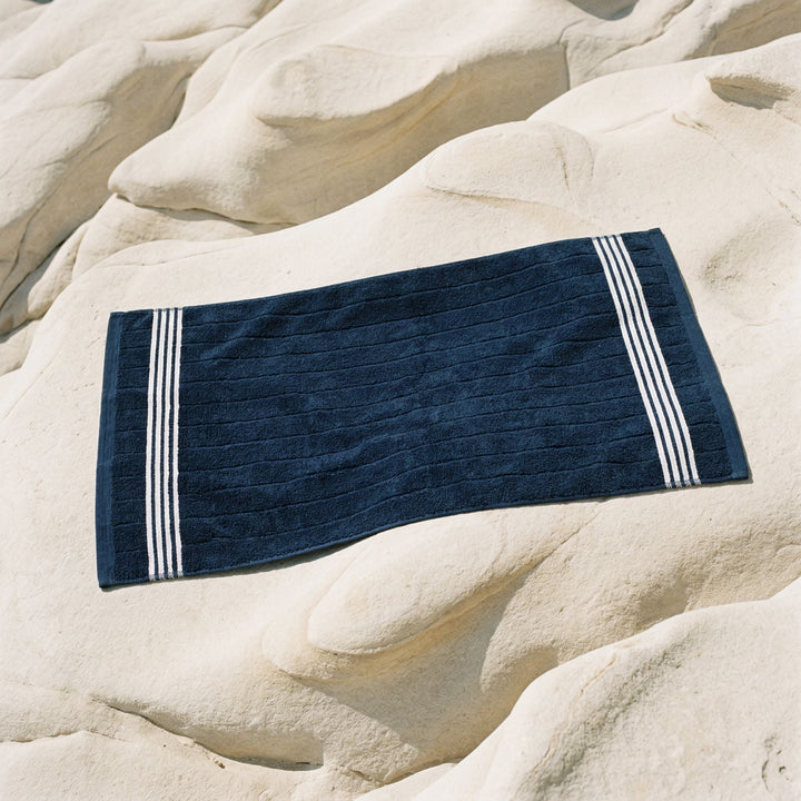 Navy cotton bath towel with white striped border, laid on rocks
