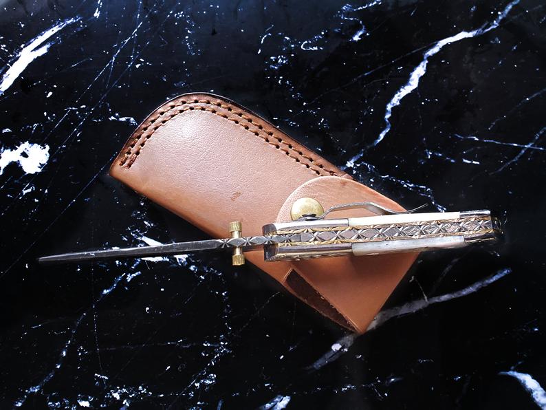Engraved folding knife closed in tan leather sheath with snap strap and damascus detailing