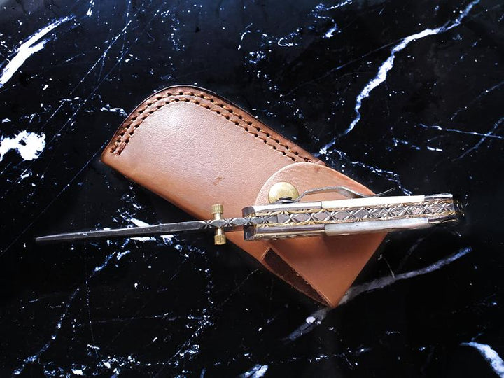 Engraved folding knife closed in tan leather sheath with snap strap and damascus detailing