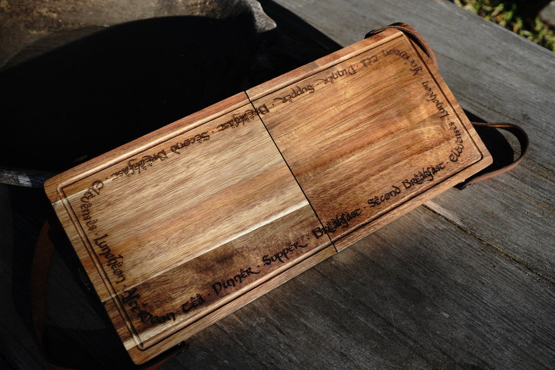 Open acacia folding cutting board with two sections and leather strap on dark surface