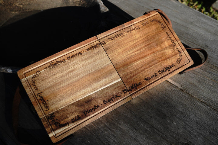Open acacia folding cutting board with two sections and leather strap on dark surface