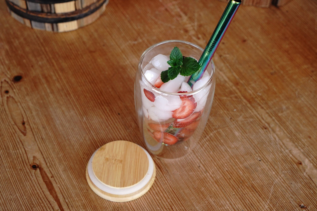 Double-walled glass mug with ice and strawberry slices; bamboo lid resting nearby.