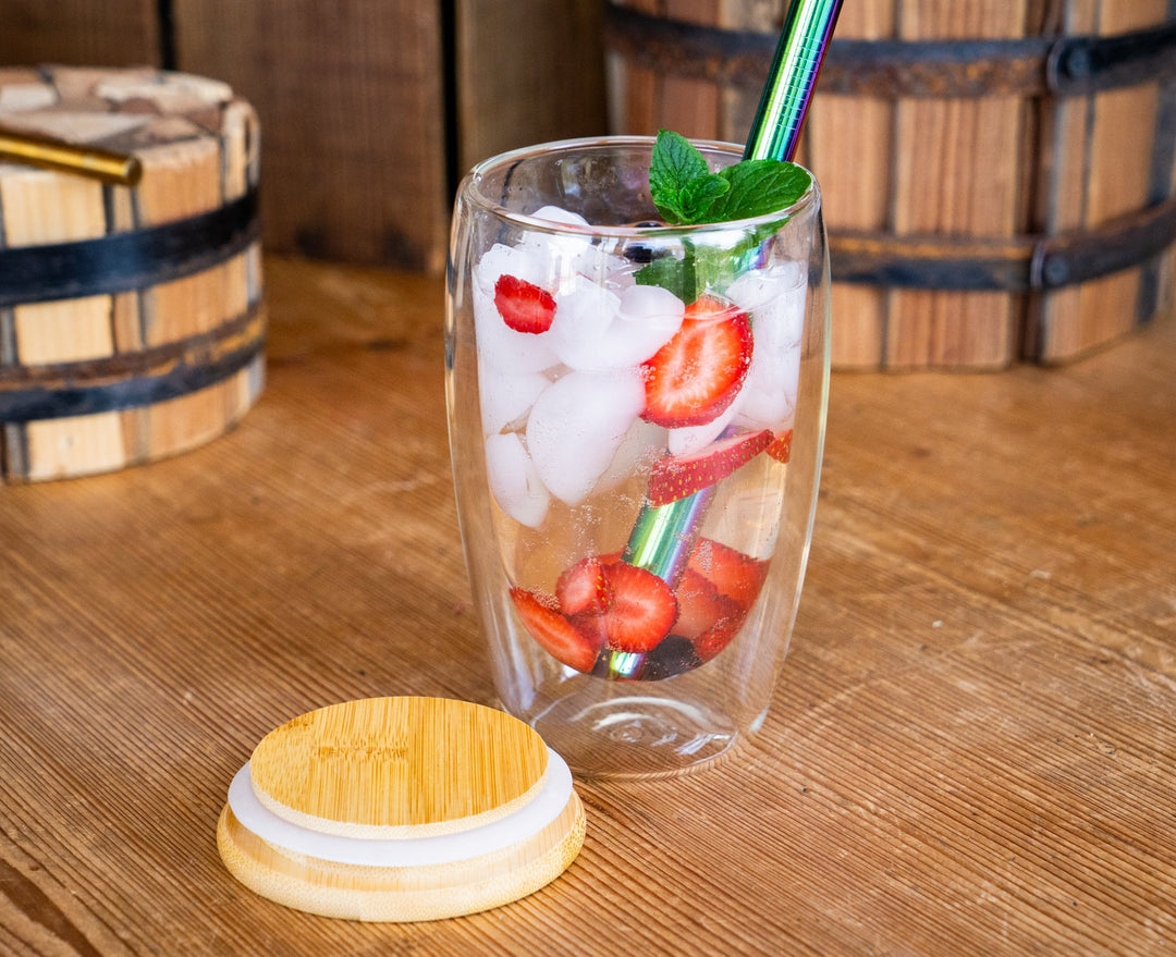 Double-walled glass mug filled with ice and strawberry slices; bamboo lid on base.