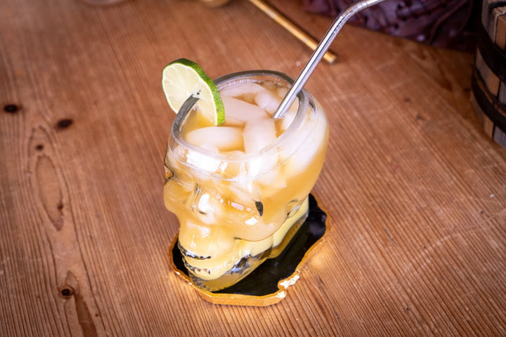Skull whiskey glass with yellow drink, lime wedge, and metal straw on coaster