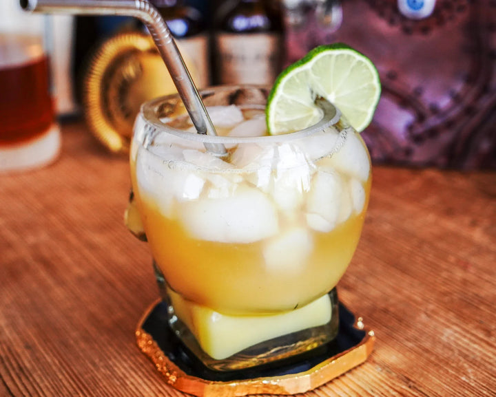 Skull whiskey glass with pale yellow drink, ice cubes, lime wedge, and straw