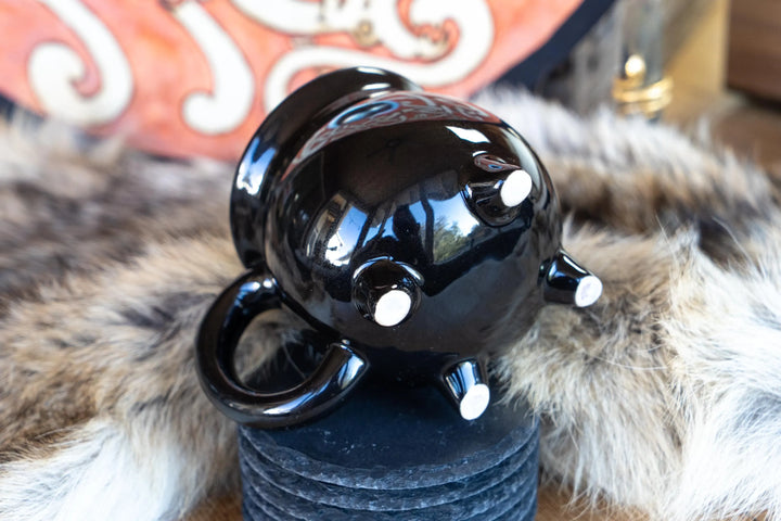 Black ceramic cauldron mug with side handle, glossy finish, three feet detail.