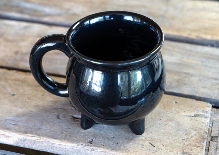 Black ceramic cauldron-style coffee mug with side handle and three legs on wood.