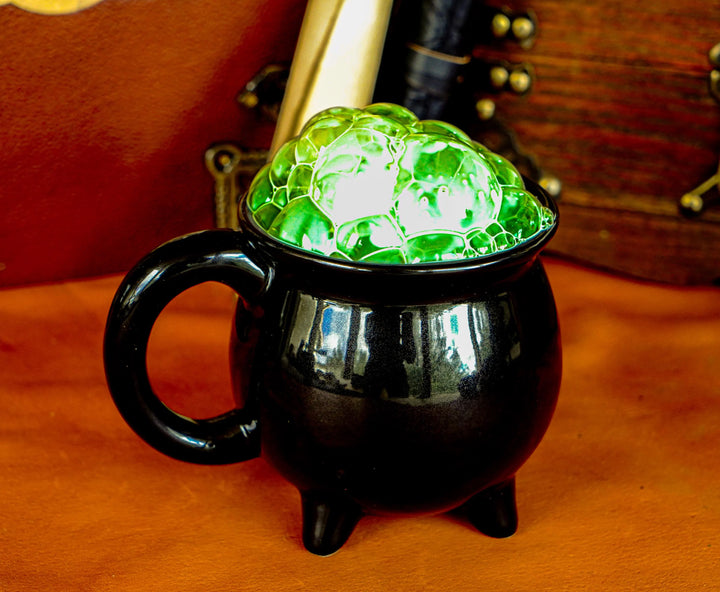 Black ceramic cauldron mug with side handle and three legs, filled with bright green ice.