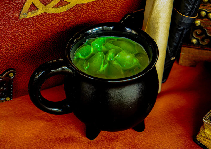 Black ceramic cauldron mug with side handle and three legs, green glowing ice close-up.