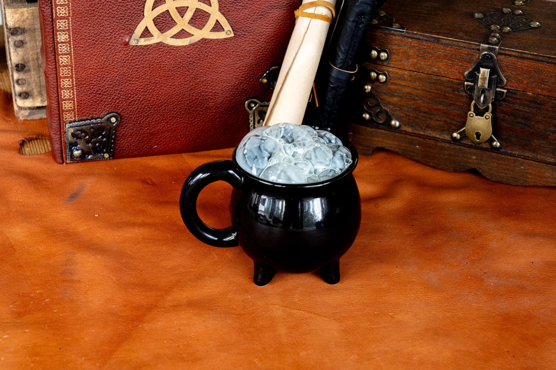 Black ceramic cauldron mug with side handle and three legs, filled with pale blue ice.