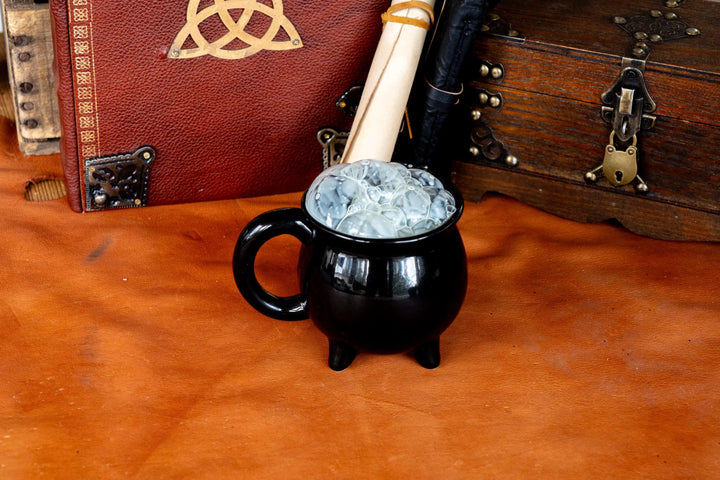 Black ceramic cauldron mug with side handle and three legs, filled with pale blue ice.