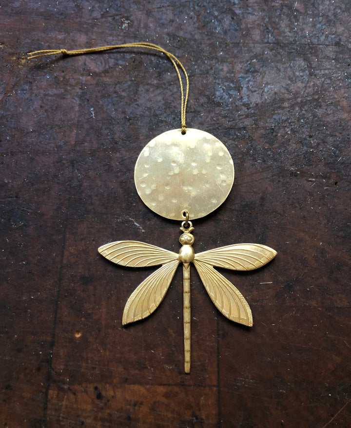 Gold-tone dragonfly ornament with round disc top and long body, suspended by a cord.