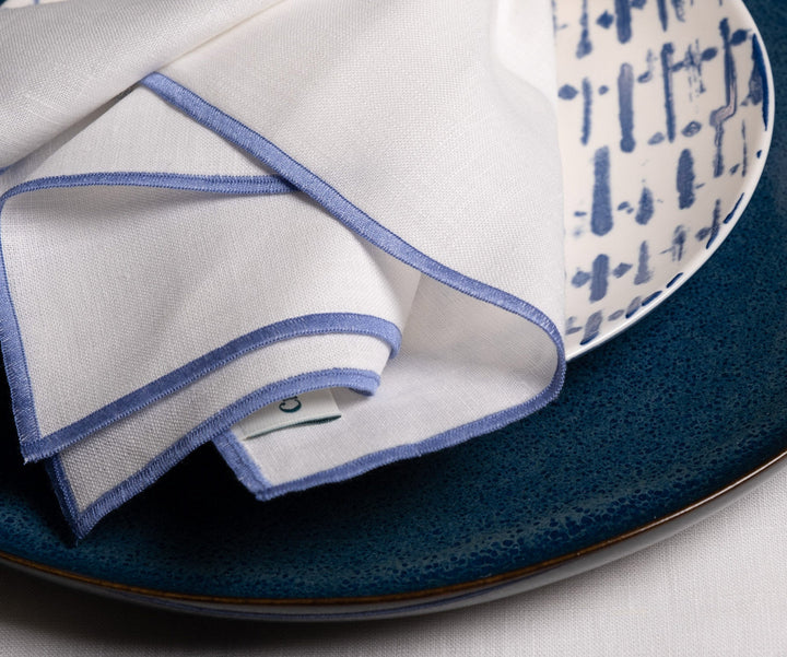 Close-up of folded white linen napkin with blue stitched border on a plate
